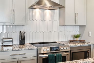 Bright modern kitchen with white vertical diamond-tile backsplash, stainless steel range hood and electric stove, granite countertops with toaster, knife block, built-in microwave and small potted plant.
