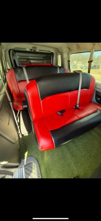 Sunlit passenger van interior with red-and-black vinyl bench seats, gray seatbelts, green carpeted floor and visible side step.