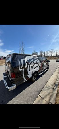 Black cargo van with a bold white-and-red bulldog mural parked at a suburban street curb beside an empty lot, leafless trees and a clear blue sky overhead.