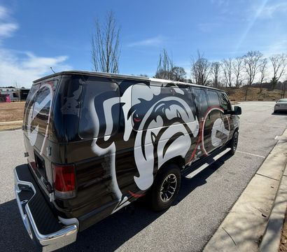 Black cargo van with a bold white-and-red bulldog mural parked at a suburban street curb beside an empty lot, leafless trees and a clear blue sky overhead.