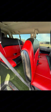 Passenger van interior with custom red-and-black leatherette bench seats, gray seatbelts, and green turf flooring, daylight visible through side windows.