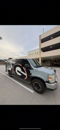 Branded black-and-gray cargo van with bold red-and-white circular logo parked in an urban multi-level parking garage lot under an overcast sky.
