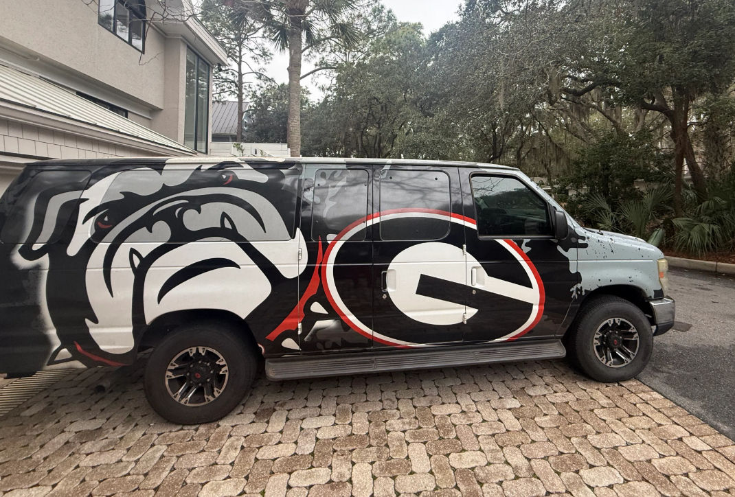 Bold black van wrapped with a snarling bulldog and oversized red-and-black 'G' emblem, parked on a brick paver driveway beside a suburban house and moss-draped oak trees.