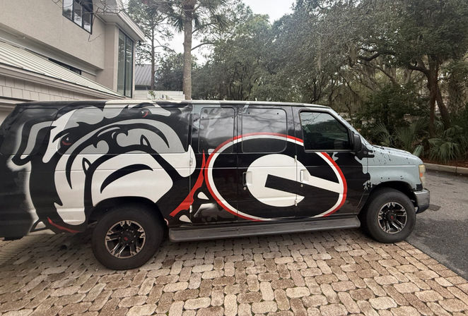Bold black van wrapped with a snarling bulldog and oversized red-and-black 'G' emblem, parked on a brick paver driveway beside a suburban house and moss-draped oak trees.