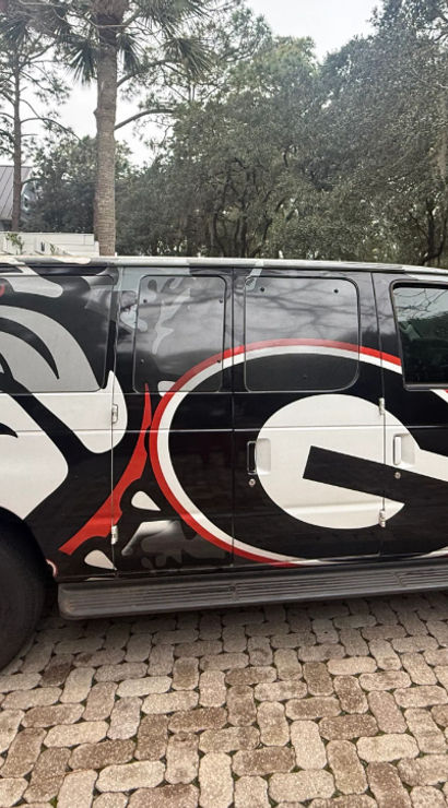 Bold black van wrapped with a snarling bulldog and oversized red-and-black 'G' emblem, parked on a brick paver driveway beside a suburban house and moss-draped oak trees.