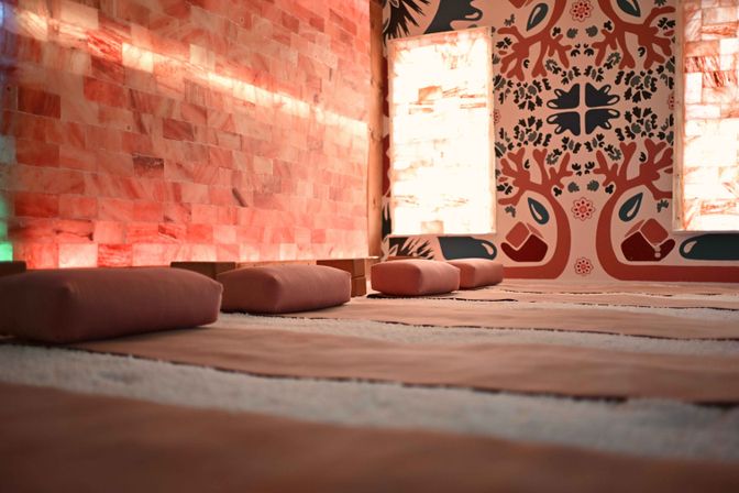 Cozy pink-hued Himalayan salt therapy room with backlit salt brick wall, decorative mural and rows of floor mats topped with meditation cushions in a serene wellness studio
