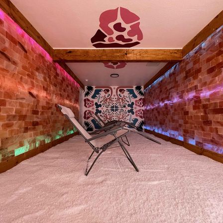 Cozy indoor salt room with two reclining lounge chairs on a white salt floor, pink Himalayan salt brick walls, LED accent lighting and a colorful floral mural.
