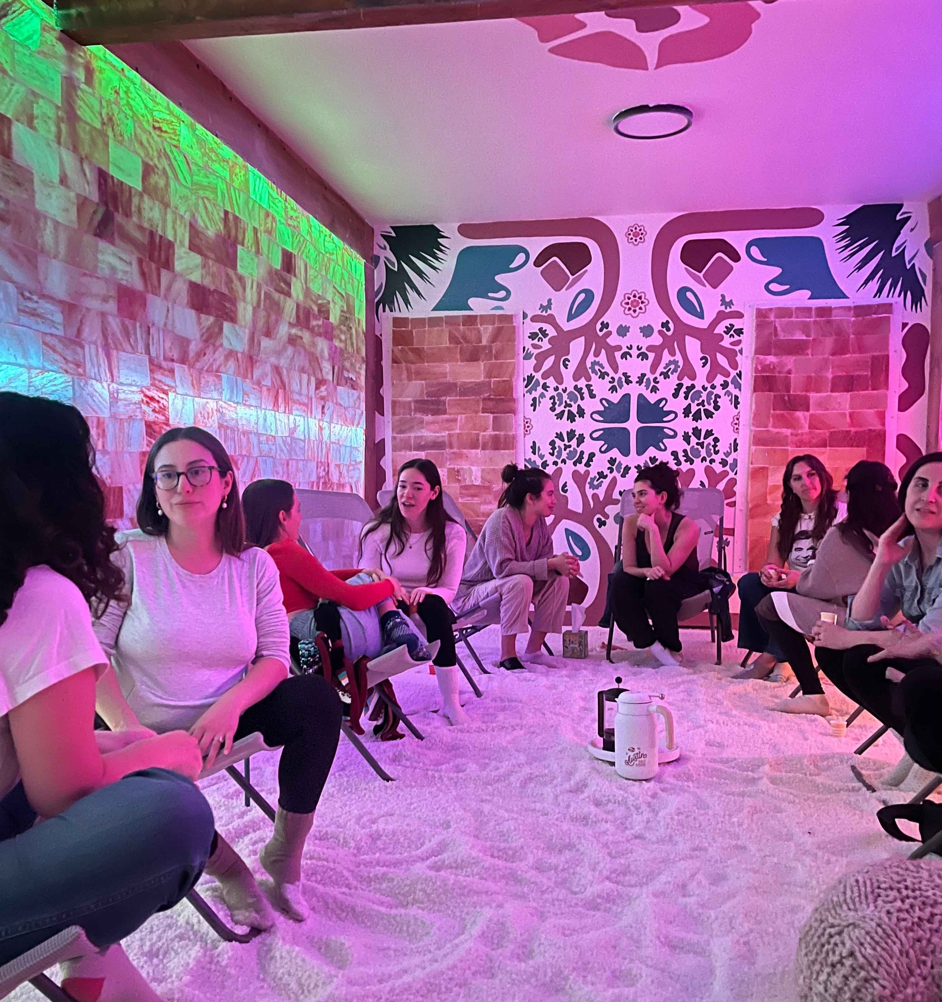 Group of women chatting and relaxing in a cozy pink- and green-lit indoor salt room, seated on folding chairs over a white salt-covered floor around a small teapot, with decorative mural and salt-block wall panels in the background.