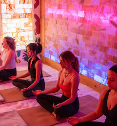 Women meditating cross-legged on yoga mats in a Himalayan salt room with glowing pink-purple backlit salt brick walls, soft salt floor and calming wellness studio ambiance.