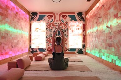 Person seated cross-legged on a mat in a glowing salt-room yoga studio, hands raised in meditation toward a colorful mural, pink salt-brick walls lit with green and pink LED accents and cushioned bolsters on a white salt-covered floor.