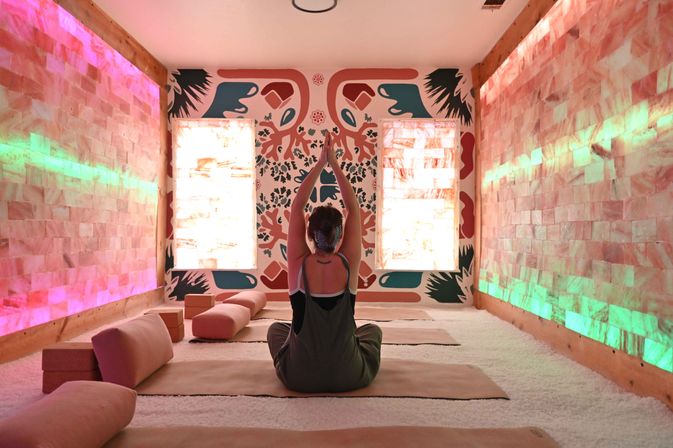 Person seated cross-legged on a mat in a glowing salt-room yoga studio, hands raised in meditation toward a colorful mural, pink salt-brick walls lit with green and pink LED accents and cushioned bolsters on a white salt-covered floor.