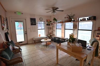 Cozy sunlit storefront lounge with wicker chairs and green cushions, patterned rug and round coffee table, wooden desk with laptop, potted plants on shelves and large front windows overlooking the street.