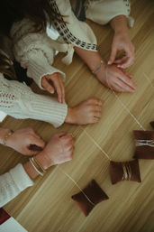 Close-up of hands in cozy sweaters arranging delicate gold necklaces and bracelets on brown velvet display pillows on a wooden table