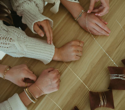 Close-up of hands in cozy sweaters arranging delicate gold necklaces and bracelets on brown velvet display pillows on a wooden table