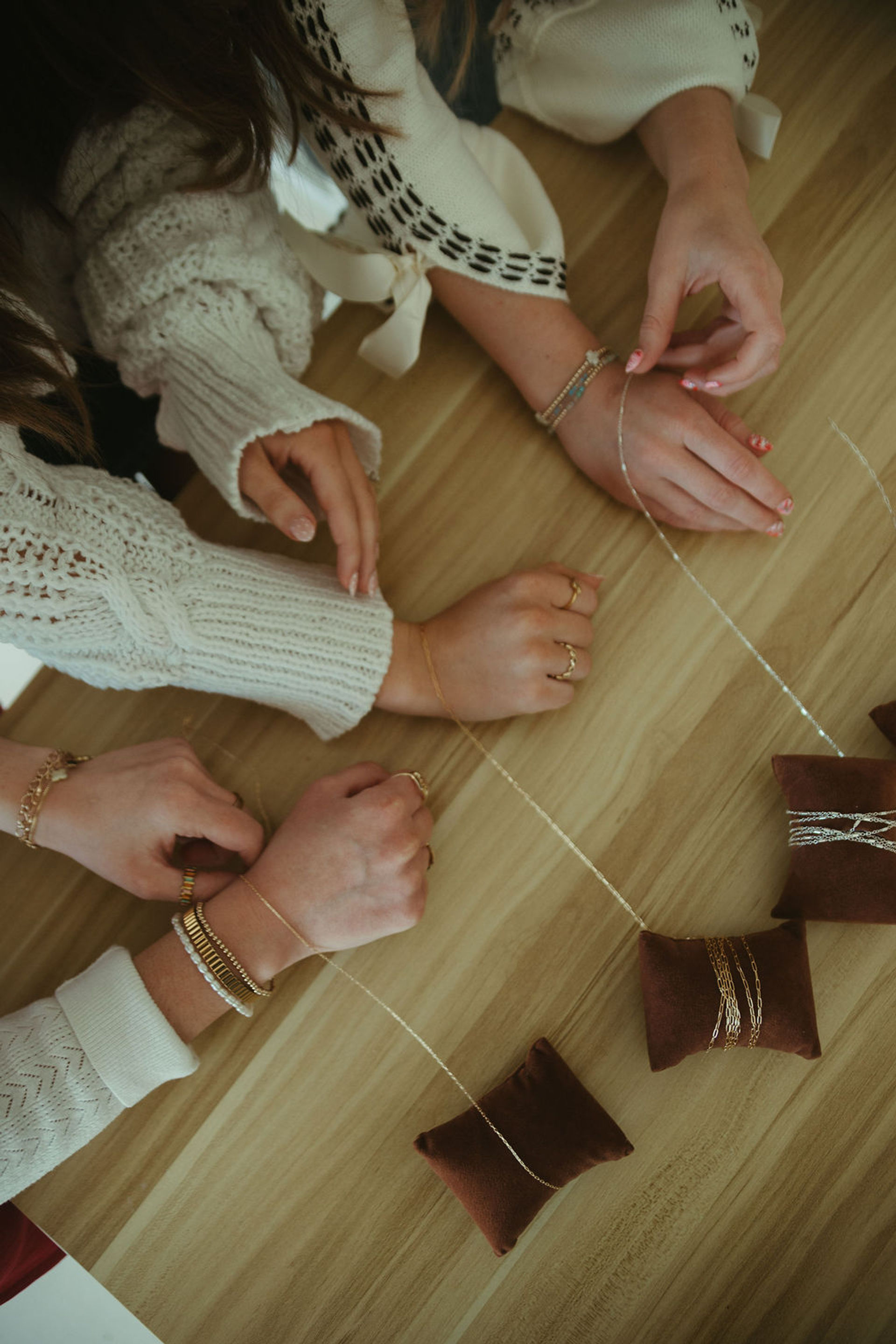 Close-up of hands in cozy sweaters arranging delicate gold necklaces and bracelets on brown velvet display pillows on a wooden table