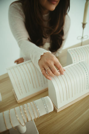 Close-up of a woman's hand wearing delicate rings selecting thin gold and silver chain necklaces from white tabletop jewelry displays on a wooden table in a bright boutique