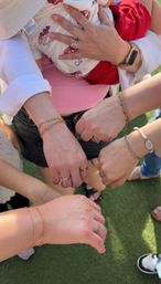 Top-down photo of several women's hands and wrists showing matching gold chain bracelets and rings, one holding a baby in red shorts, gathered on a green lawn — outdoor family/friends moment.