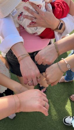 Top-down photo of several women's hands and wrists showing matching gold chain bracelets and rings, one holding a baby in red shorts, gathered on a green lawn — outdoor family/friends moment.