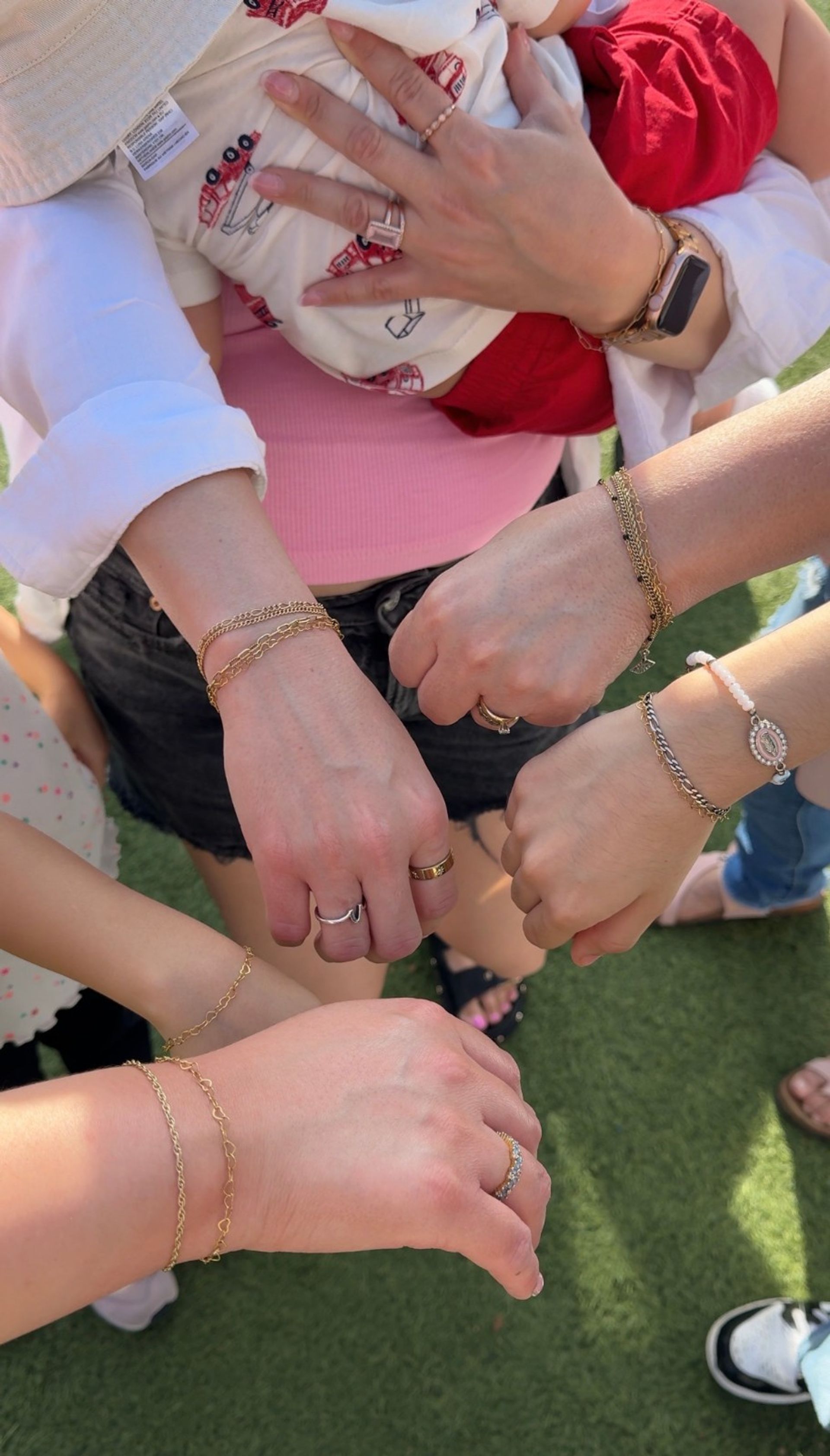 Top-down photo of several women's hands and wrists showing matching gold chain bracelets and rings, one holding a baby in red shorts, gathered on a green lawn — outdoor family/friends moment.
