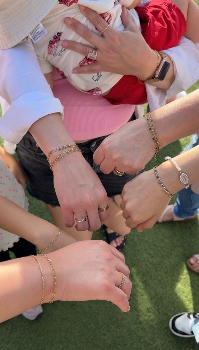 Top-down photo of several women's hands and wrists showing matching gold chain bracelets and rings, one holding a baby in red shorts, gathered on a green lawn — outdoor family/friends moment.