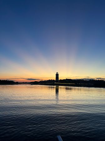 Silhouetted coastal lighthouse at sunset with golden rays fanning across a twilight sky, reflected on calm rippling harbor water.