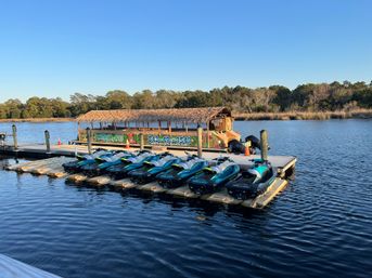 Row of turquoise-and-black jet skis lined up on floating docks beside a thatched-roof rental boat at a calm coastal marina with marsh grasses and trees under a clear blue sky.