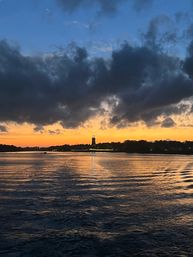 Coastal lighthouse silhouette at sunset with a golden horizon, dramatic dark clouds overhead and shimmering reflections on rippling bay water.