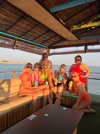 Group of seven women in colorful swimsuits and sunglasses enjoying a sunset pontoon boat cruise on coastal waters, smiling and holding drinks with a sandy shoreline in the background.
