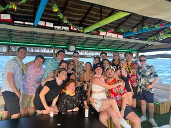 Smiling group of friends in Hawaiian shirts and leis posing on a covered tiki-style party boat over blue ocean water, festive decorations and a captain hat for a fun boat celebration