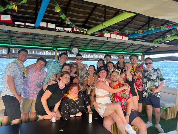 Smiling group of friends in Hawaiian shirts and leis posing on a covered tiki-style party boat over blue ocean water, festive decorations and a captain hat for a fun boat celebration