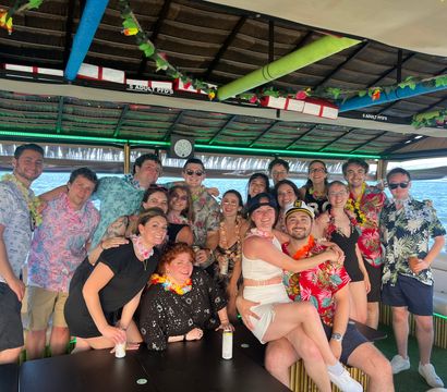 Smiling group of friends in Hawaiian shirts and leis posing on a covered tiki-style party boat over blue ocean water, festive decorations and a captain hat for a fun boat celebration