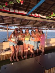 Group of six adults wearing leis and summer outfits smiling and posing on a tiki-style party boat at sunset with calm coastal waters visible through open sides
