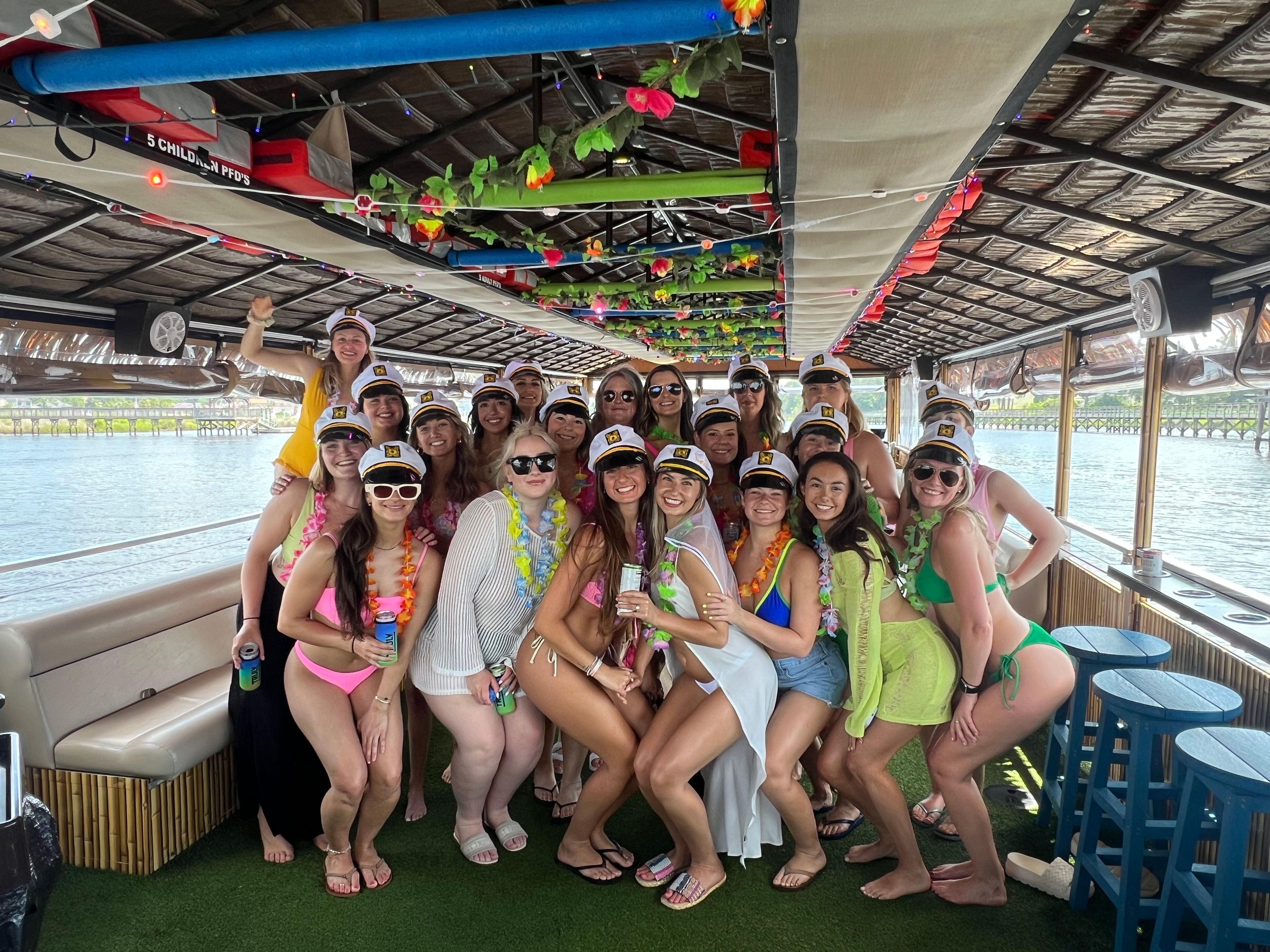 Group of women in swimsuits wearing captain hats and colorful leis posing on a decorated tiki-style party boat during a sunny waterfront cruise