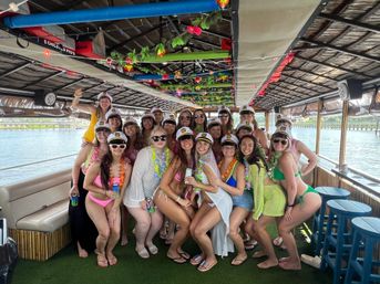 Group of women in swimsuits wearing captain hats and colorful leis posing on a decorated tiki-style party boat during a sunny waterfront cruise