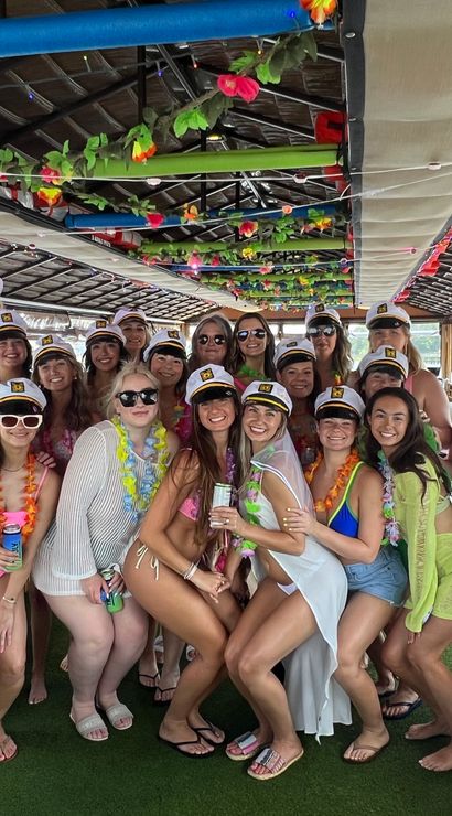 Group of women in swimsuits wearing captain hats and colorful leis posing on a decorated tiki-style party boat during a sunny waterfront cruise