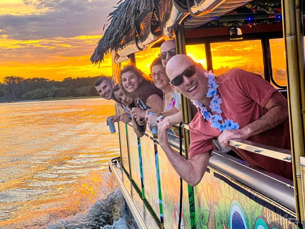 Group of smiling adults leaning out of a colorful tiki-style boat with leis and drinks on a sunset river cruise, golden water reflecting the vibrant sky and distant tree-lined shore.