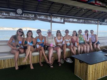 Group of people in swimsuits sitting on a tiki-style party boat by a sandy shoreline, smiling and holding drinks under a shaded deck on a sunny day.