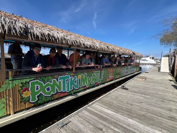 Tiki-style party boat with a thatched roof docked at a sunny coastal marina, a group wearing leis leaning on the railing alongside a wooden dock and yachts in the background