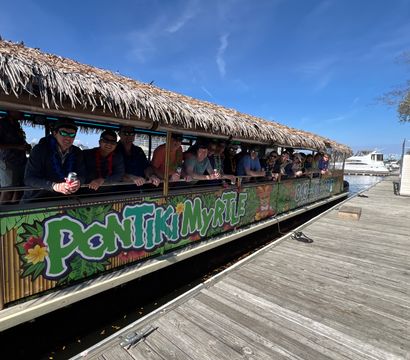 Tiki-style party boat with a thatched roof docked at a sunny coastal marina, a group wearing leis leaning on the railing alongside a wooden dock and yachts in the background
