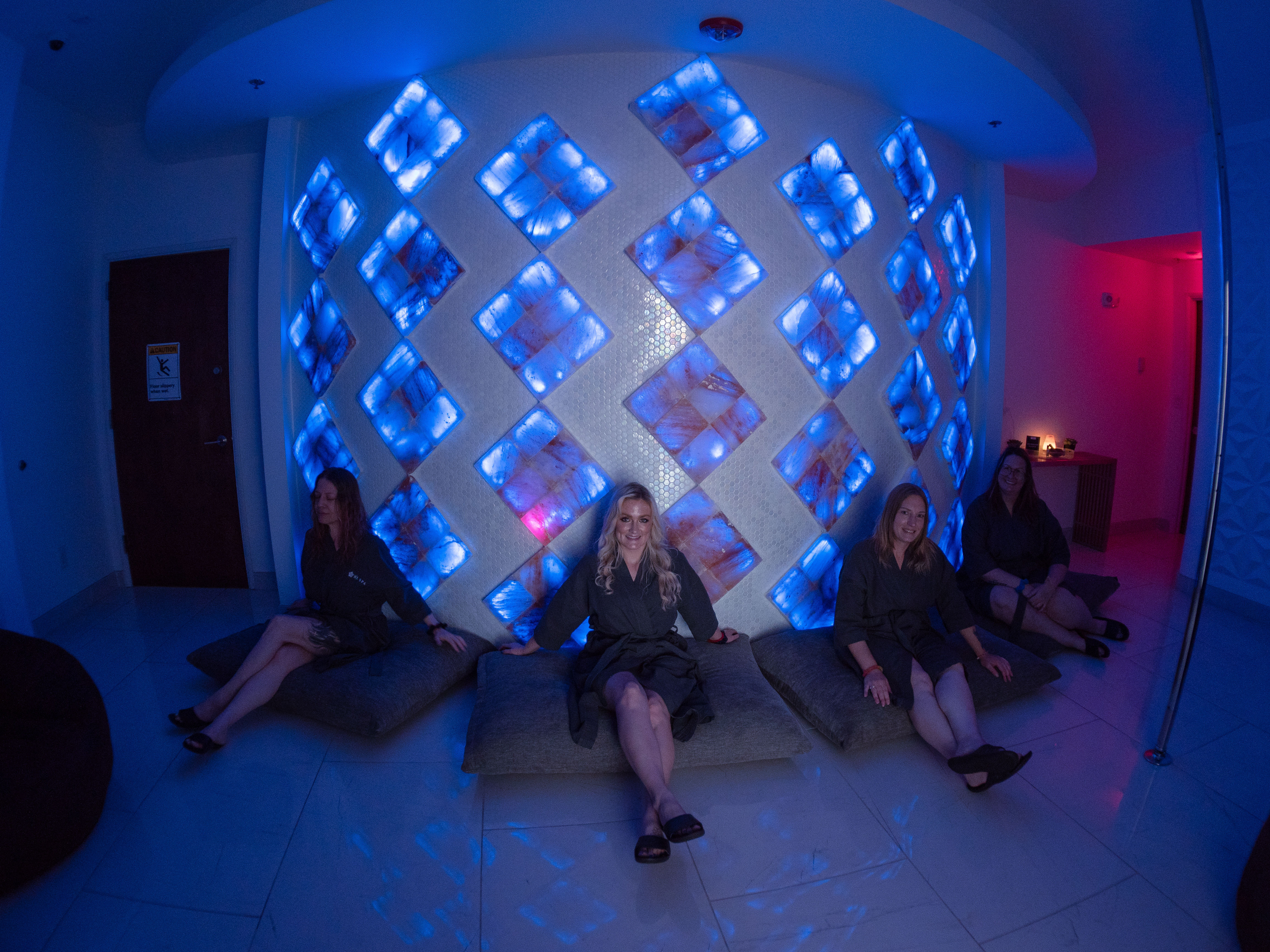Four women in black spa robes relax on floor cushions in a wellness lounge in front of a curved wall of LED‑lit blue salt blocks arranged in a diamond pattern, creating a moody, relaxing atmosphere.