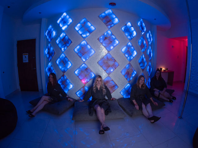 Four women in black spa robes relax on floor cushions in a wellness lounge in front of a curved wall of LED‑lit blue salt blocks arranged in a diamond pattern, creating a moody, relaxing atmosphere.