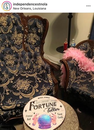 Pair of vintage carved-wood armchairs in blue-and-gold damask with a pink feather boa; round "Fortune Teller" sign showing a crystal ball on a small stone table — New Orleans parlor vibe.