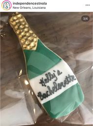 Green and gold champagne bottle sugar cookie decorated with a white label bearing a bachelorette message, wrapped in clear plastic on a wooden table