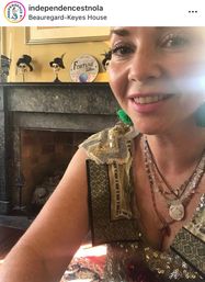 Selfie of a smiling person with glittery eye makeup, green tassel earring and layered necklaces, wearing a sequin-patterned top; behind them a marble fireplace mantel displays three decorative skulls in hats and a round "Fortune Teller" sign in a historic parlor.
