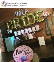 Bridal shower setup in New Orleans with gold foil 'BRIDE' and silver 'NOLA' balloons above a row of gift bags and favors, a veil-draped chair, floral gift bag, and a vintage-style 'Fortune Teller' sign with a crystal ball.