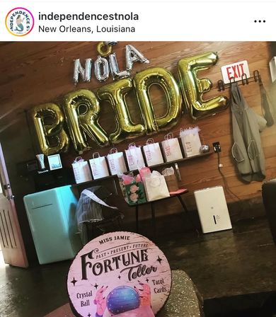 Bridal shower setup in New Orleans with gold foil 'BRIDE' and silver 'NOLA' balloons above a row of gift bags and favors, a veil-draped chair, floral gift bag, and a vintage-style 'Fortune Teller' sign with a crystal ball.