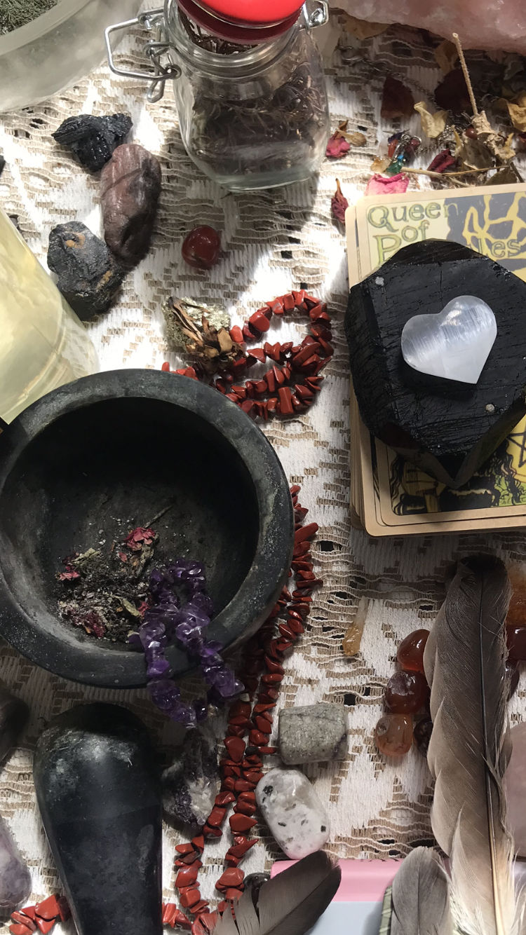 Boho spiritual altar setup with crystals and tarot: black mortar holding dried herbs and amethyst chips, red jasper chip necklace, tarot cards topped by a heart-shaped selenite, glass jar and feathers on a lace cloth.