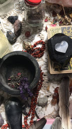 Boho spiritual altar setup with crystals and tarot: black mortar holding dried herbs and amethyst chips, red jasper chip necklace, tarot cards topped by a heart-shaped selenite, glass jar and feathers on a lace cloth.