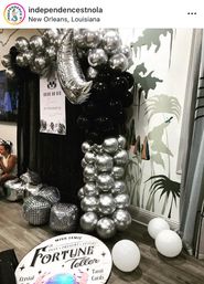 Bachelorette party setup in New Orleans with black and silver balloon column topped by a metallic crescent moon, mirrored disco-ball cubes, white floor balloons, and a round “Fortune Teller” sign against a tropical mural backdrop.