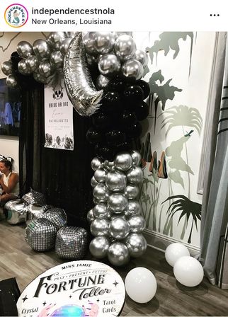 Bachelorette party setup in New Orleans with black and silver balloon column topped by a metallic crescent moon, mirrored disco-ball cubes, white floor balloons, and a round “Fortune Teller” sign against a tropical mural backdrop.